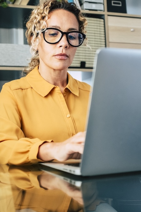Professional businesswoman wearing eyeglasses and a yellow shirt, concentrating while typing on a laptop computer, reflecting modern office work and digital communication
